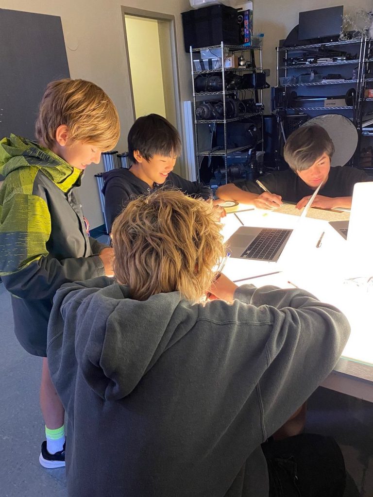 Four boys smiling and laughing who are gathered around a lightbox sketching drawings and using digital technology